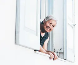 Uma mulher idosa e sorridente está à janela a espreitar para a rua, num dia de verão. Crédito: Mike Jones (Canva) 