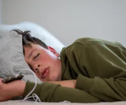 um adolescente dorme com o telemóvel na mão, sob um fundo neutro. Crédito: Canva.