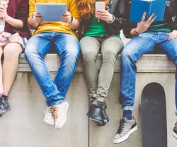 vários adolescentes estão a ver conteúdos no telemóvel e ipad, sentados num muro, num ambiente citadino. Crédito: Canva