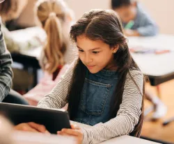 Uma menina de cabelos escuros, com um tablet na mão, tendo como fundo uma sala de aula. Crédito: Shutterstock
