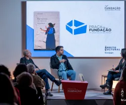 Uma plateia, de costas, a olhar para um palco, onde estão dois homens sentados a falar.