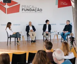 Quatro pessoas em palco, sentadas em cadeiras pretas, a falar com plateia a assistir. Logo e texto "Praça da Fundação".