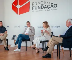 Quatro pessoas sentadas em cadeiras pretas, a debater entre si. Ao fundo, o texto "Praça da Fundação"