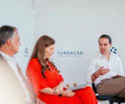 Três pessoas sentadas num palco. Dois homens de fato, uma senhora com um vestido vermelho. Ao fundo, o logotipo da Fundação.