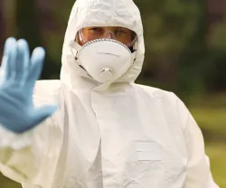 Imagem de uma pessoa, vestida com um fato de segurança branco, máscara, óculos e luvas azuis.