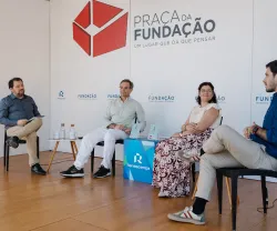 Quatro pessoas sentadas num palco, em cadeiras pretas, a apresentar um livro que está pousado em cima de uma mesa. Fundo com logotipos da Fundação.
