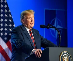 Donald Trump a falar, num púlpito. Fundo azul, com uma bandeira dos EUA e o símbolo da Nato. Créditos: Gints Ivuskans/shutterstock
