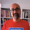 Imagem de um homem, o analista Bobby Gosh, vestido com uma t-shirt vermelha, com uma estante de livros atrás.