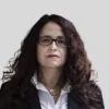 Imagem de uma mulher, vestida com uma camisa branca e um casaco preto, com um colar ao pescoço.