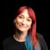 Retrato a cores da comunicadora Ana Markl, de cabelo vermelho e azul, vestida com uma t-shirt preta.
