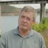 Retrato de um homem, de óculos, vestido com uma camisa. Uma barragem de fundo.
