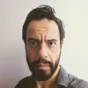 Retrato a cores de um homem, de barba e cabelo escuro, vestido com uma camisa cinzenta, sobre uma parede branca.