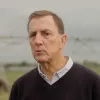 Retrato de um homem vestido com uma camisa e uma camisola por cima. Ambiente de campo.