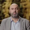 Retrato de um homem, careca e com barba, vestido com uma camisa azul clara. Uma pintura cobre o fundo.