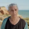 Retrato de uma senhora de cabelo branco, vestida com uma t-shirt preta. Ao fundo, o mar.
