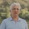 Retrato a cores de um homem de cabelo branco, com óculo, e vestido com um polo azul claro. Um rio e vegetação cobrem o fundo.