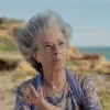 Retrato a cores de uma senhora idosa, que usa óculos e um lenço ao pescoço. Uma praia cobre o fundo.