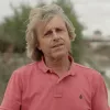 Retrato a cores de um homem com cabelo claro e pelos ombros, vestido com um polo rosa. 
