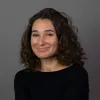 Retrato a cores de uma mulher, sorridente, de cabelo ondulado e escuro, vestida com uma camisola preta. Fundo cinzento escuro.