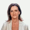 Retrato de uma mulher, sorridente, de cabelo castanho, vestida com um top branco e um blazer bege com riscas pretas. DR