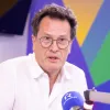 Um homem de óculos, num estúdio de rádio, a falar para um microfone. Fundo com várias cores.