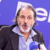 Retrato a cores de um homem com barba e cabelo grisalhos, vestido com uma camisa azul e um blazer preto, a falar para um microfone.