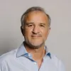 Retrato a cores de um homem, de cabelo grisalho, expressão sorridente. Veste uma camisa azul clara. DR