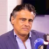 Um homem, vestido com um fato azul escuro e uma camisa azul clara, num estúdio de rádio, com um microfone à frente. Créditos: Fábio Oliveira.