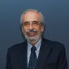 Retrato a cores de um homem de óculos, cabelo e barba grisalho, vestido de fato. Fundo azul.