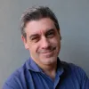 Retrato a cores de um homem, sorridente, de barba e cabelo grisalhos, vestido com uma camisa azul. Fundo cinzento.