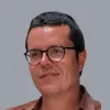 Retrato a cores de um homem, de óculos e cabelo escuro e curto, a sorrir. Fundo cinzento.