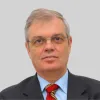 Retrato a cores de um homem de óculos e cabelo grisalho, vestido com um fato preto, uma camisa azul e uma gravata vermelha e verde. D/R
