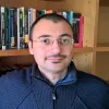 Retrato a cores de um homem que usa óculos e veste um camisola azul. Uma estante com livros ao fundo.