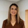 Retrato a cores de uma mulher sorridente, que veste um casaco castanho e usa um colar ao pescoço.