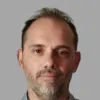 Retrato a cores de um homem de cabelo escuro e barba grisalha. Fundo cinzento. D/R