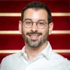 Retrato a cores de um homem com barba e de óculos, a sorrir, vestido com uma camisa branca. Fundo vermelho com riscas brancas. 