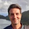 Retrato a cores de um homem a sorrir, com um lago e montanhas ao fundo.