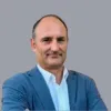 Retrato a cores de um homem vestido com uma camisa branca e um blazer azul. Fundo cinzento.D/R