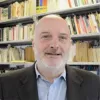 Retrato a cores de um homem com barba branca. Uma estante de livros cobre o fundo. D/R