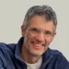 Retrato a cores de um homem que usa óculos e tem o cabelo grisalho. Veste uma camisola azul.