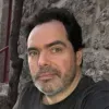 Retrato a cores de um homem com barba grisalha e cabelo escuro. Veste uma t-shirt preta. Fundo de uma parede em pedra.