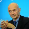 Retrato a cores de um homem, Pascal Lamy, com as mãos cruzadas. Veste um fato preto e uma camisa azul. Fundo azul claro.