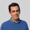 Retrato a cores de um homem a sorrir, que usa óculos e tem cabelo escuro. Veste uma camisola azul. Fundo cinzento.