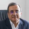 Retrato a cores de um homem com óculos, casaco preto e camisa branca, sentado numa cadeira de escritório. D/R